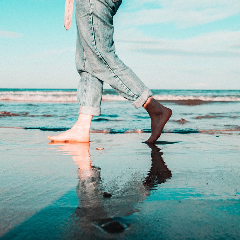 Casually dressed woman with rolled-up jeans walking barefoot on a sandy beach, capturing a serene coastal moment from the waist down